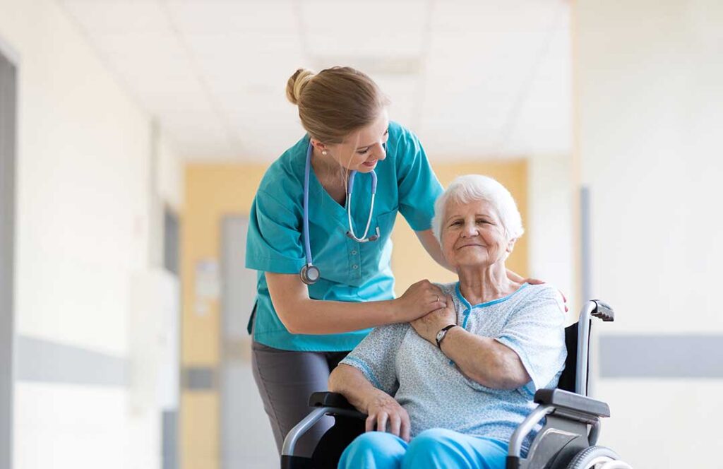 Caregiver helps an older resident walk through an assisted living hallway while another staff member observes.